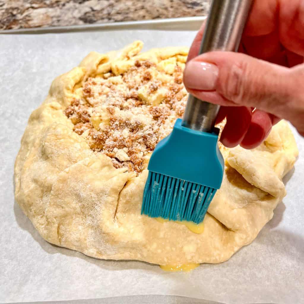 Using a silicone brush to brush egg wash on the crust of an apple Streusel galette.