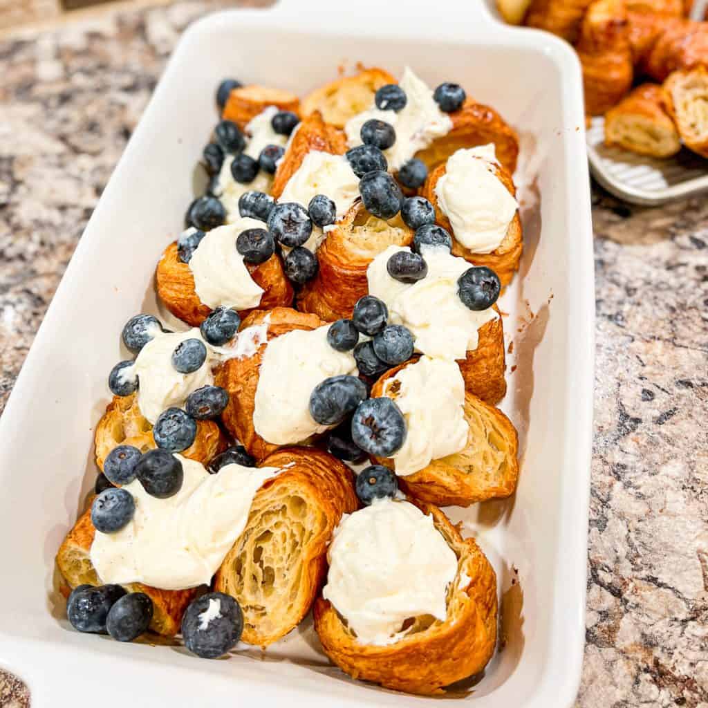 A white casserole dish containing croissants covered in blueberries and cheesecake mix.