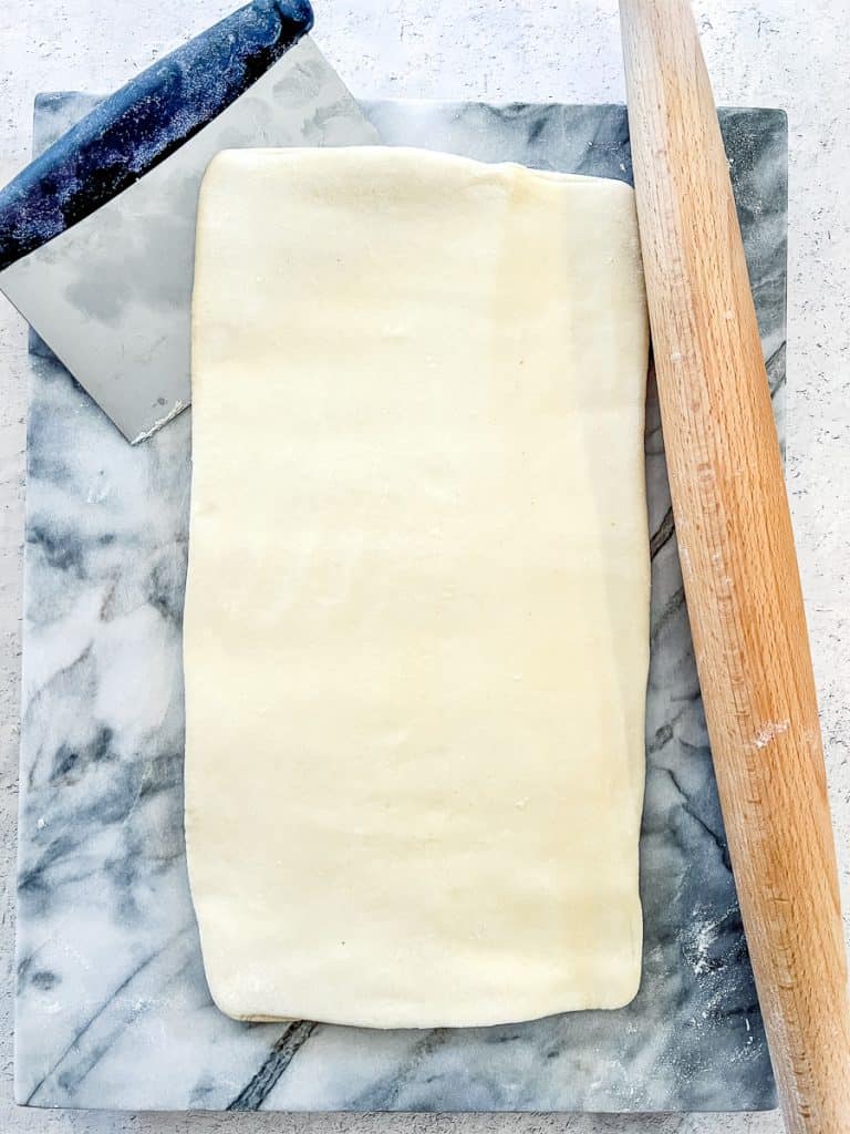 Rolled out Simple Rough Puff Pastry, after the sixth and final series of folds, on a marble board with a bench scraper and rolling pin nearby.